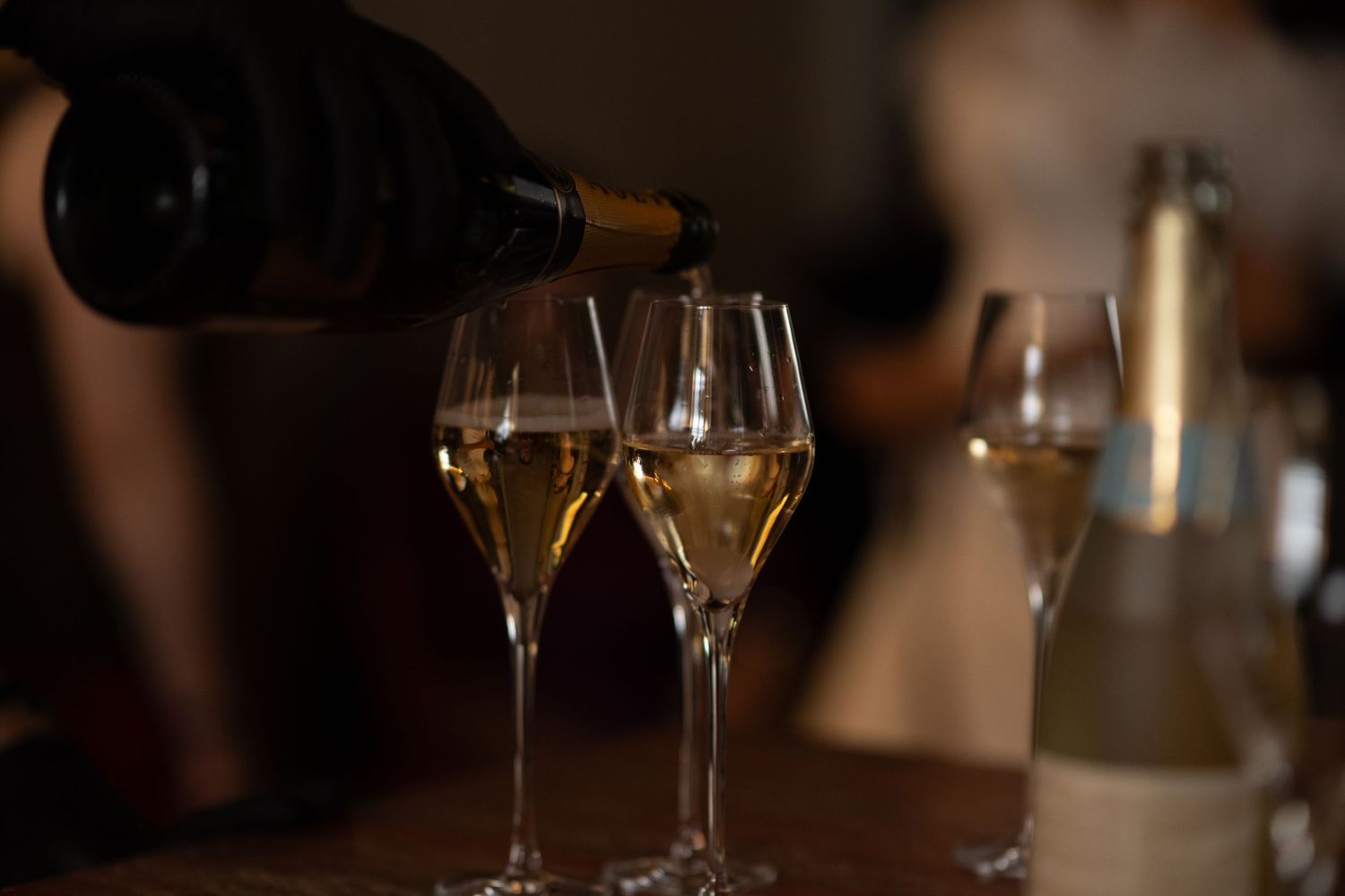 Sparkling wine being poured from a dark bottle into elegant champagne flutes on a wooden table, with a blurred bottle and guests behind.