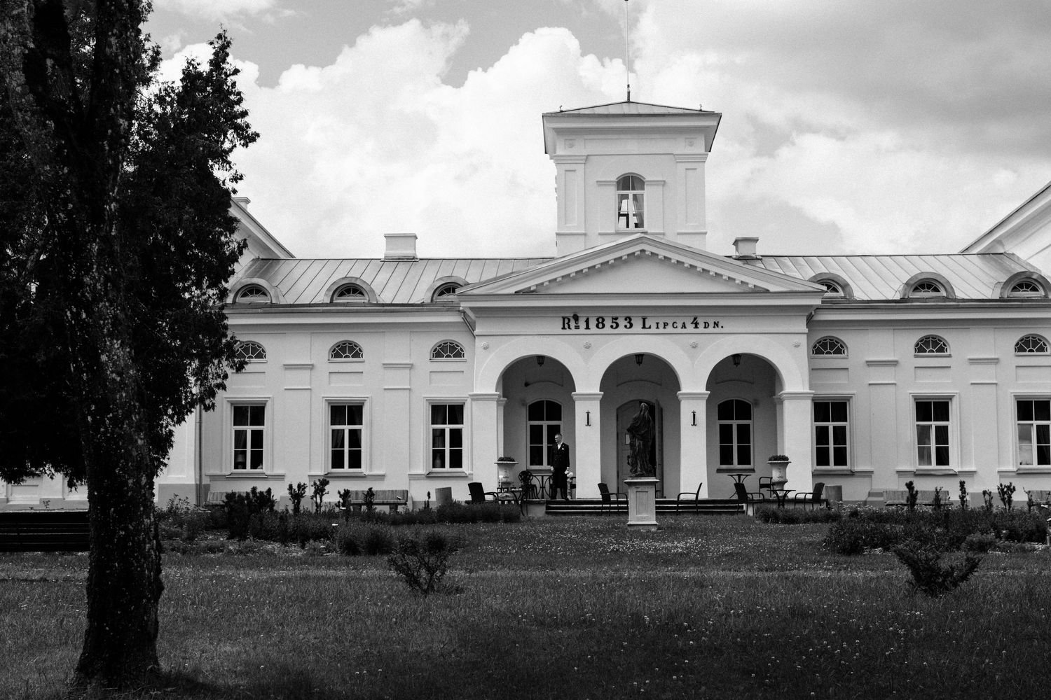 Historic white manor house with a central tower, arched entrance portico and symmetrical windows, set behind a lawn under a cloudy sky.