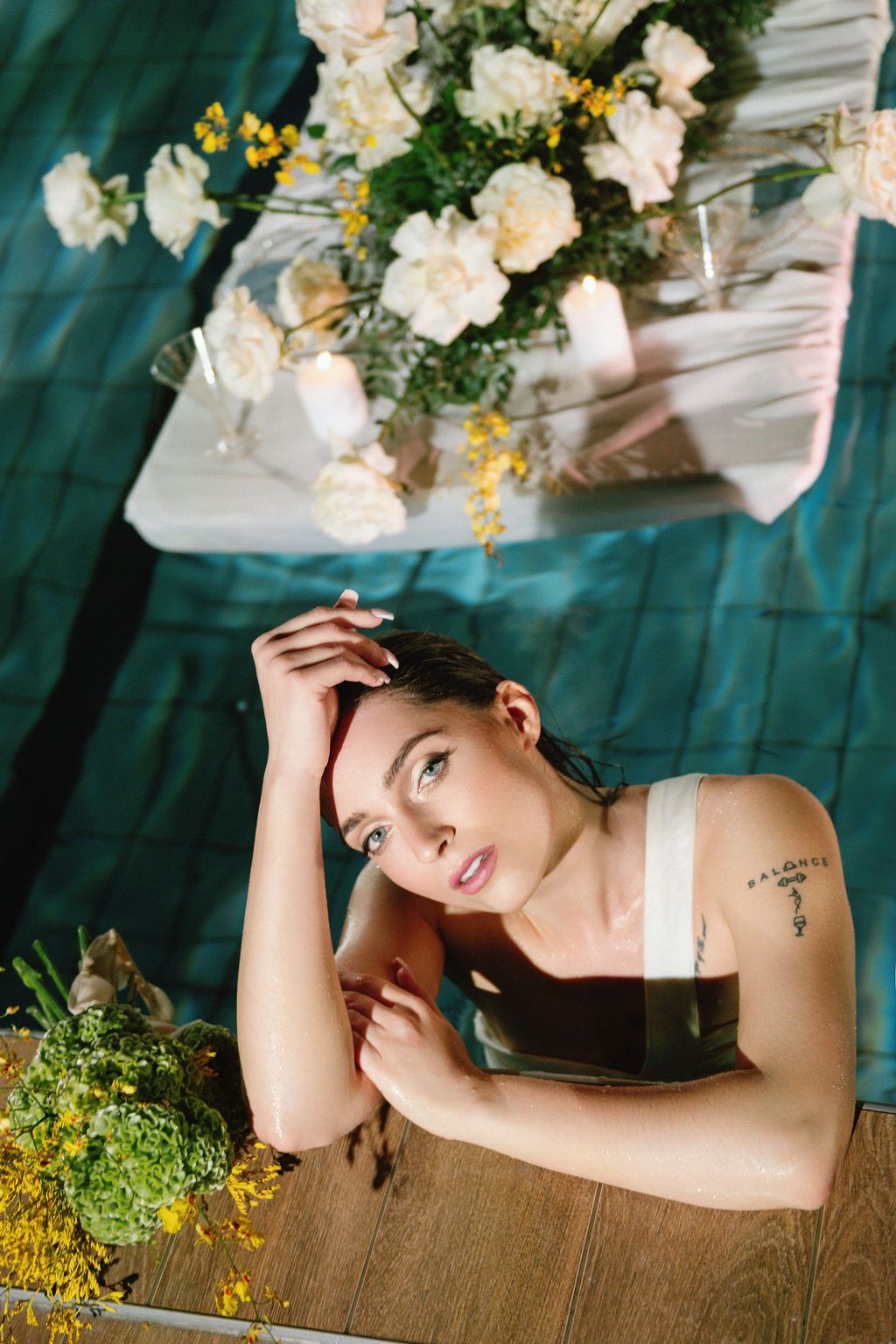 Young woman with tattoo poses near floral arrangement with white roses and yellow flowers on green backdrop.