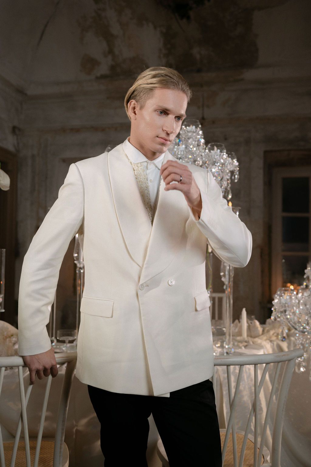 Man in ivory double-breasted tuxedo jacket with ornate bow tie poses in elegant vintage ballroom setting.