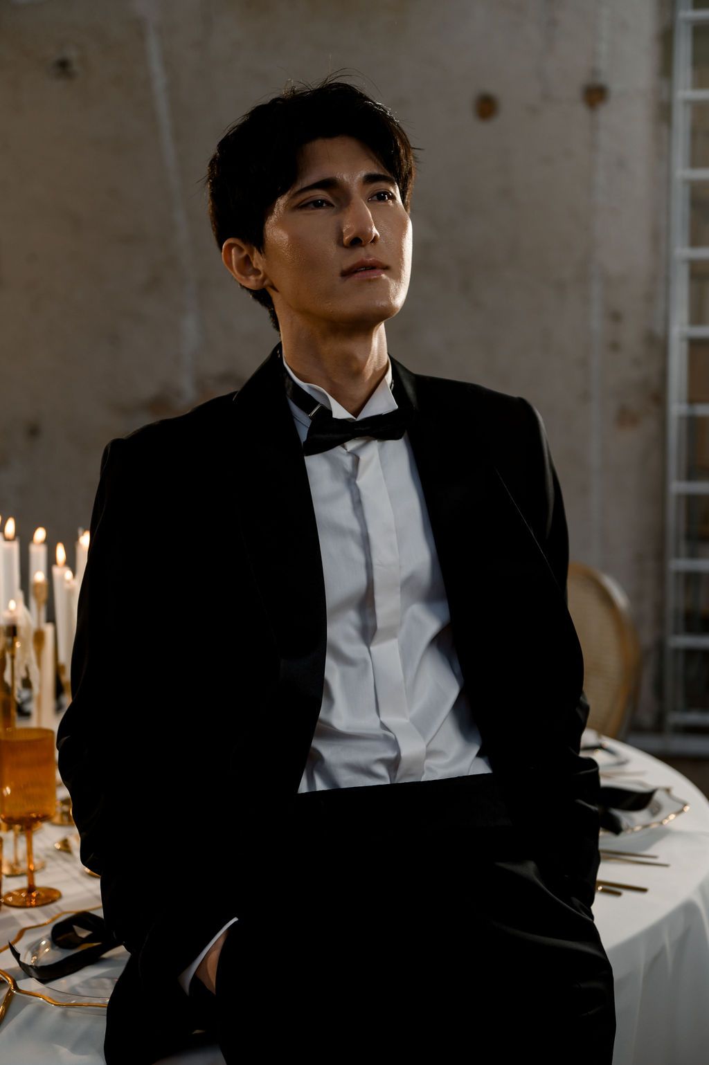 Stylish man in black tuxedo with bow tie sitting near candlelit table in moody atmospheric setting.