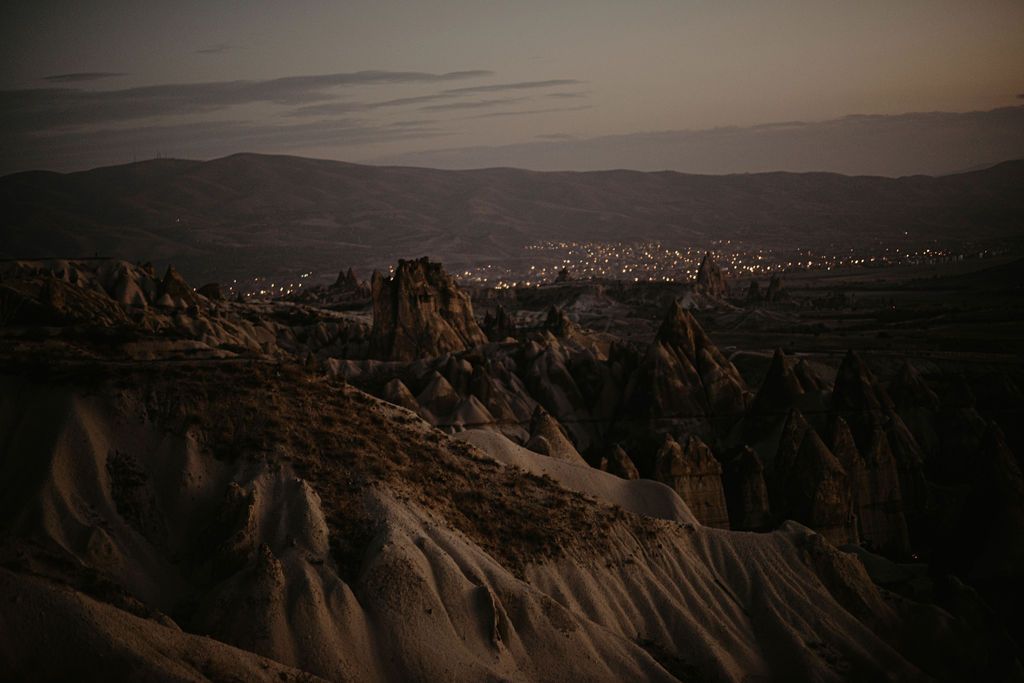 Dramatic twilight view of Cappadocia's iconic fairy chimney rock formations with a glowing town in the valley below.