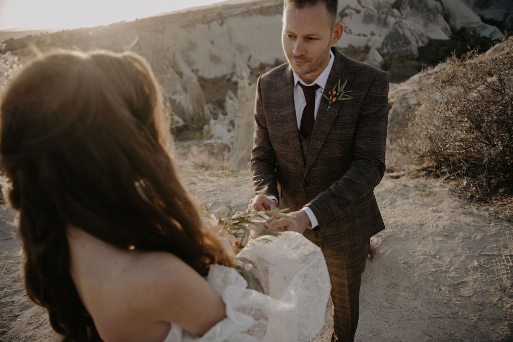 Groom in brown plaid suit holds bride's hand outdoors during golden hour wedding ceremony in rocky landscape.