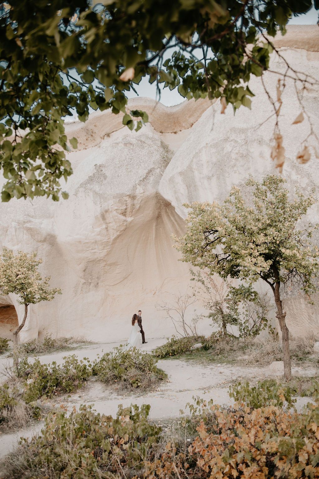 A couple embraces near dramatic white rock formations in Cappadocia, Turkey, surrounded by sparse trees and desert shrubs.
