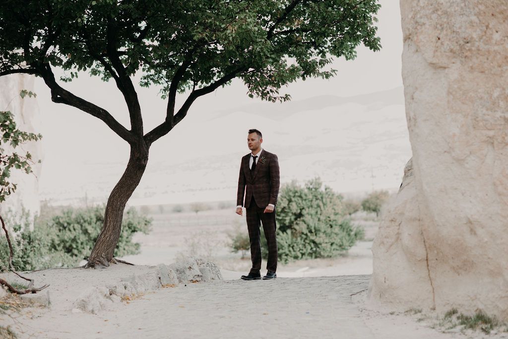 A groom in a dark suit stands between a tree and stone wall in a desert landscape.