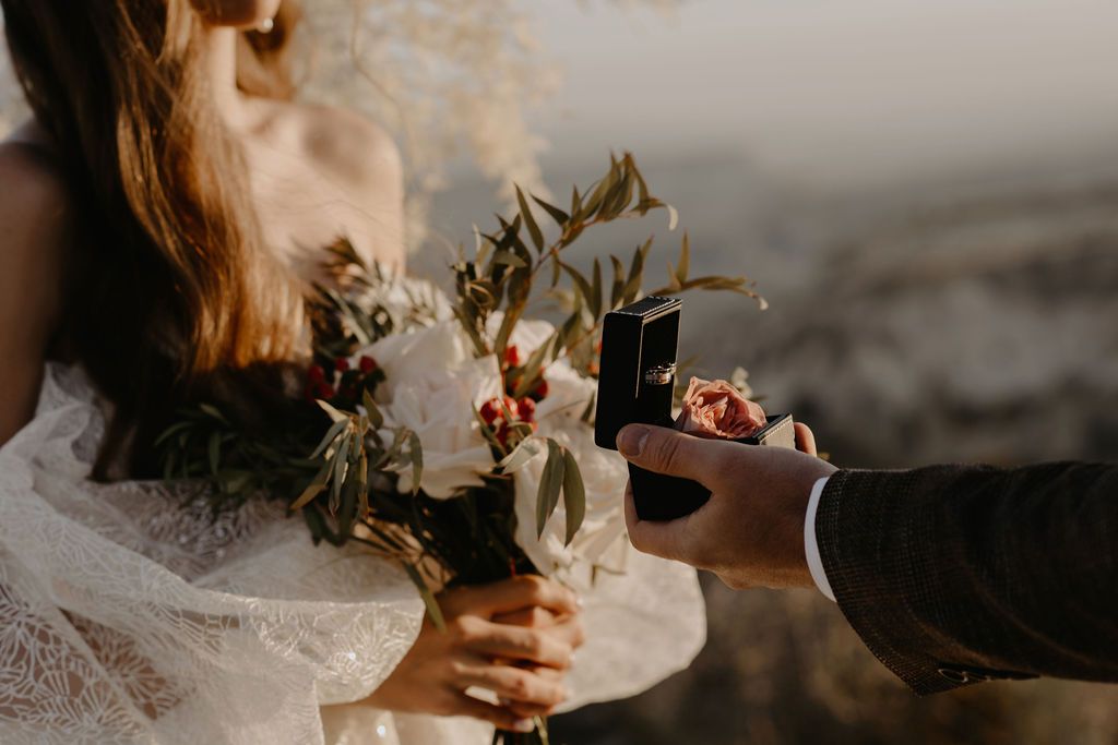 Bride holding a rustic bouquet with olive branches while groom presents a ring box during an outdoor wedding ceremony.