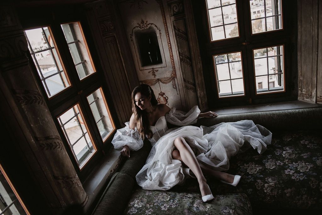 Bride in white wedding dress sitting elegantly by vintage windows in an ornate, dimly lit historic room.