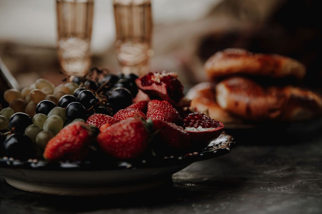 A dark plate with fresh strawberries, grapes, and pomegranate beside croissants and glasses in soft focus.
