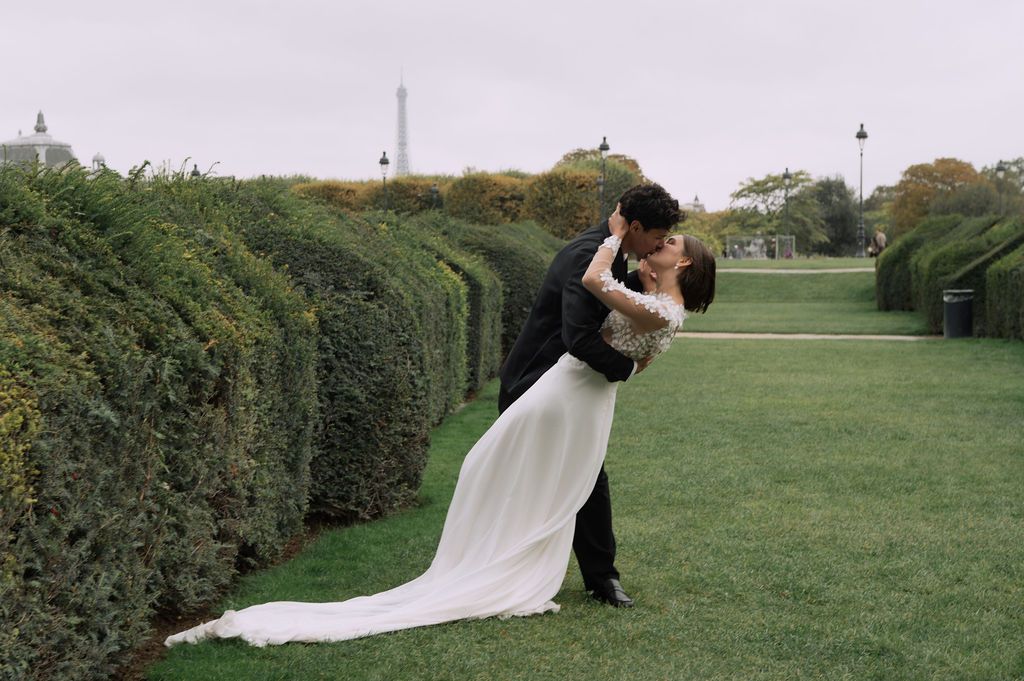 Couple sharing a romantic dip kiss in a lush green garden with the Eiffel Tower in the background on their wedding day.