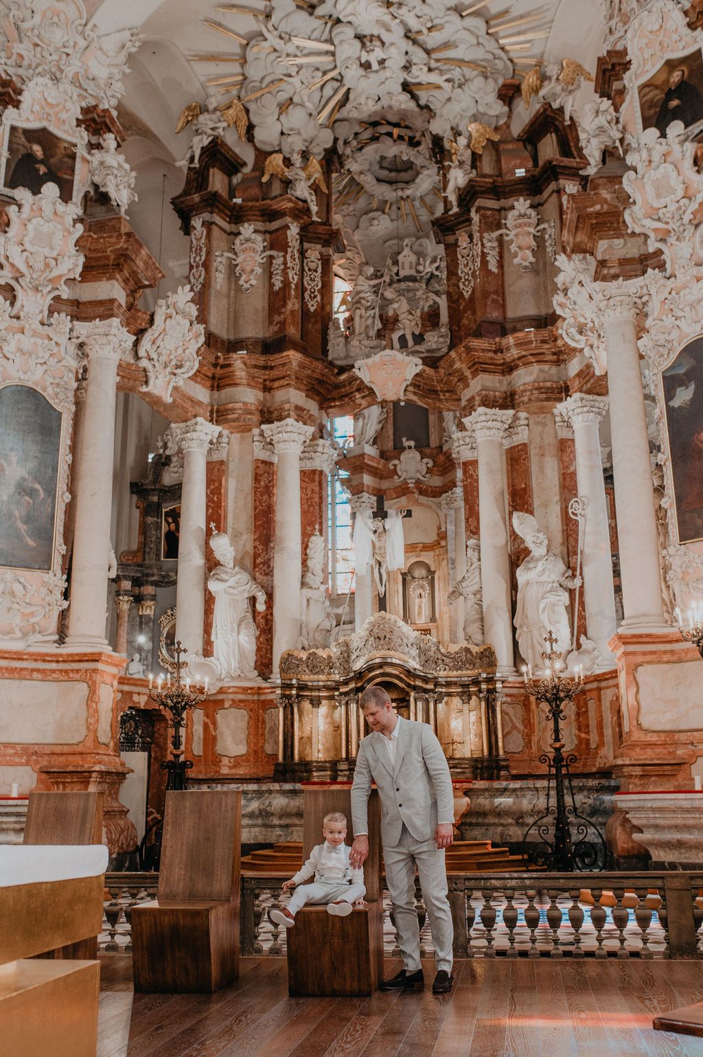 Adult and child stand before ornate Baroque church altar with elaborate white and gold stucco decorations and marble columns.