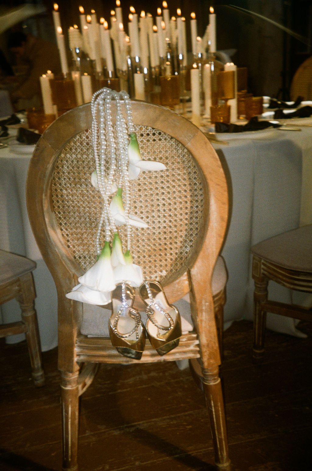 Elegant wedding chair with pearl necklace, white lily, and gold heels, set against candlelit reception table.