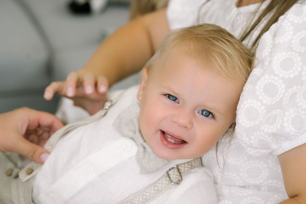 An adorable smiling baby with blue eyes sits on a womans lap wearing a white outfit at a special event.