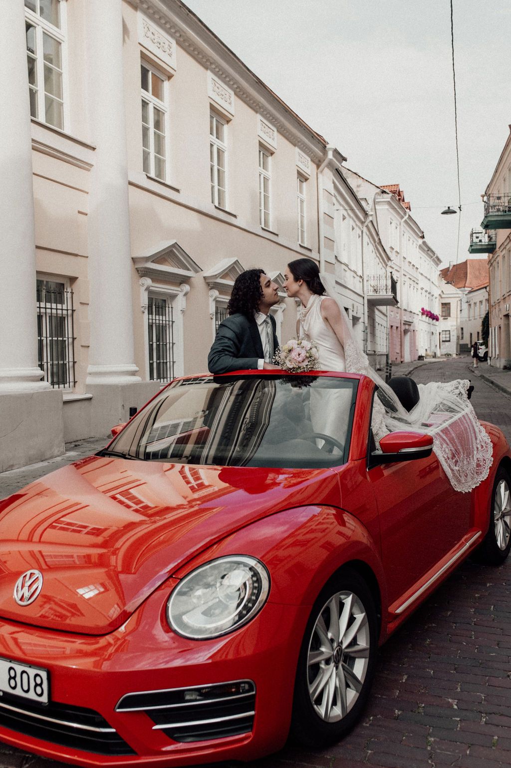 Bride and groom kissing atop a red VW Beetle convertible on a cobblestone street in a European city.