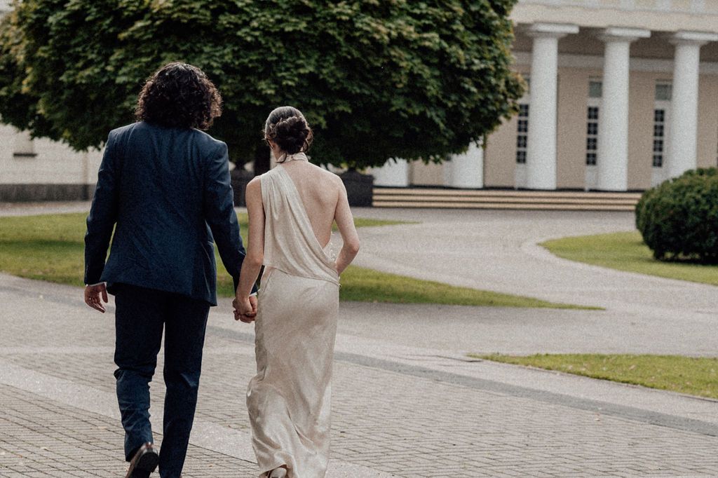 A couple in wedding attire walks hand in hand along a brick path toward a grand white columned building.