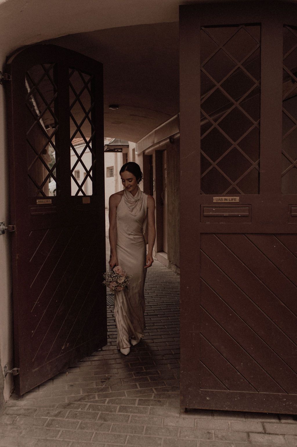 Bride in elegant satin gown holding bouquet walking through rustic wooden doors.