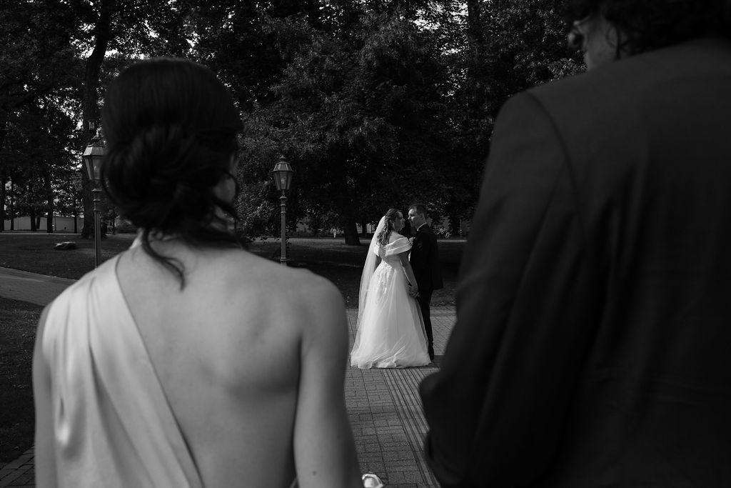 Black and white wedding photo of bride with elegant updo viewed from behind watching couple embrace in park.