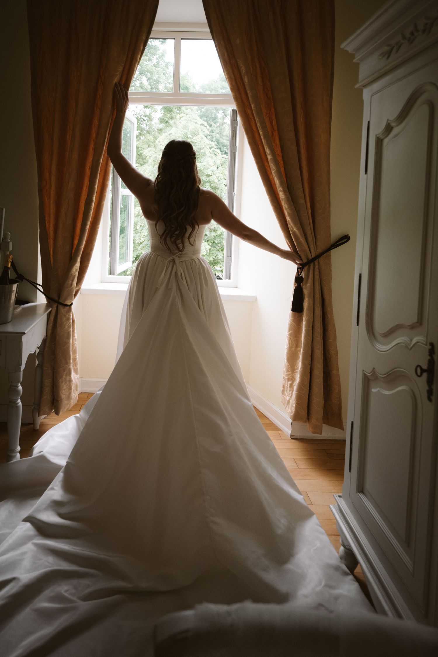 Bride in white wedding gown standing at window with golden curtains in elegant room.