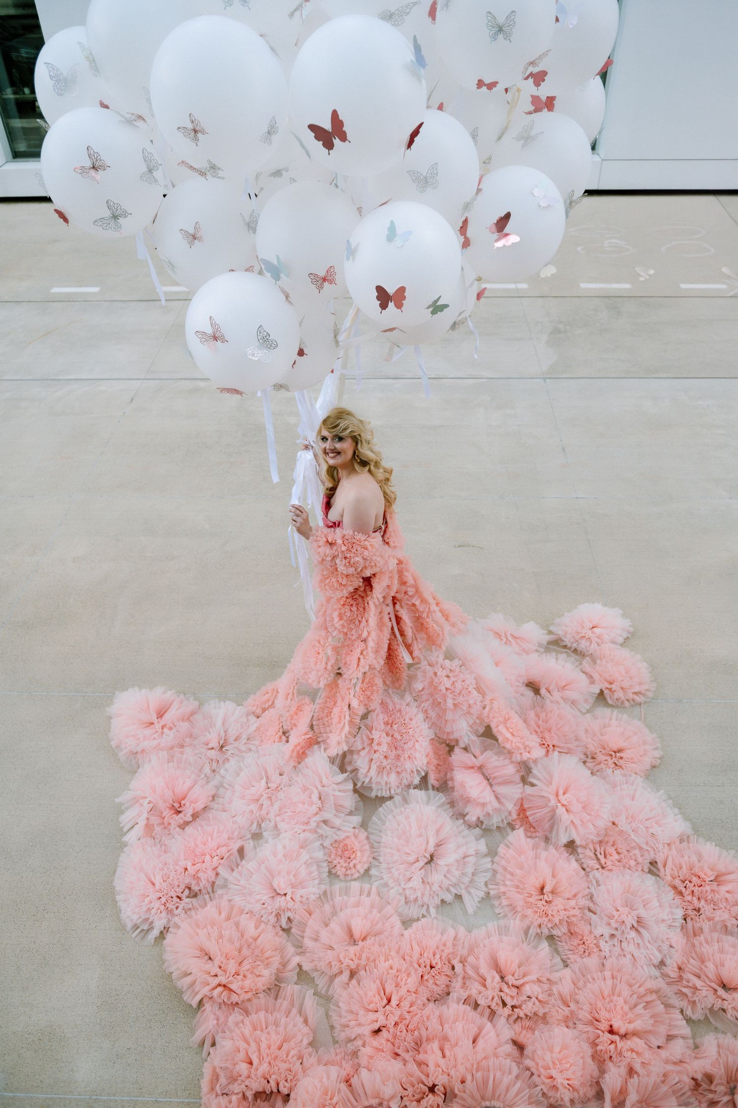 Woman in pink ruffled gown holding white butterfly balloons, elegant floral train spread on floor.