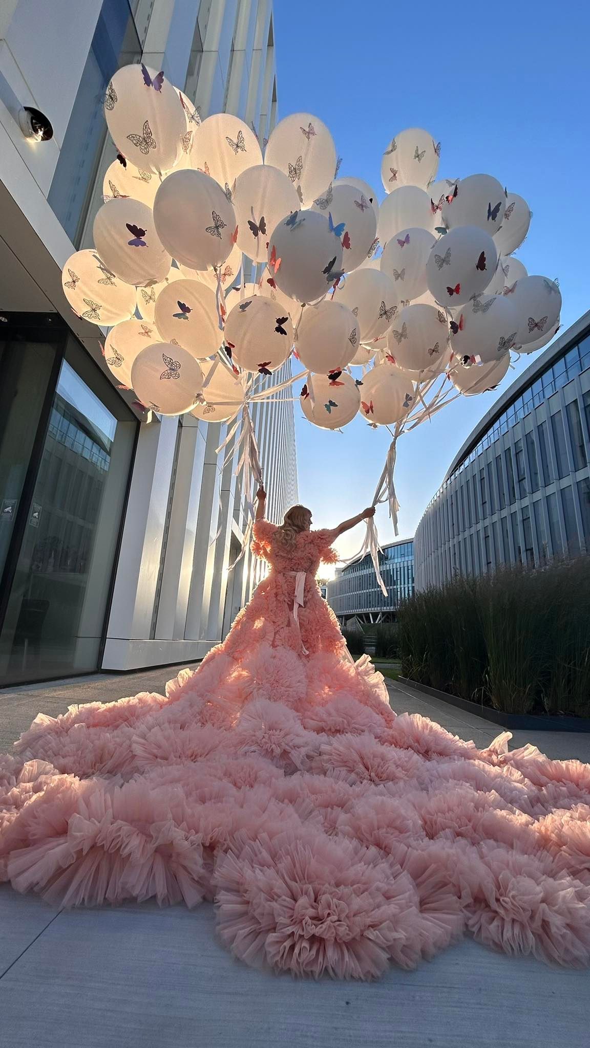 Woman in pink ruffled ballgown holds white butterfly balloons outdoors near modern building under blue sky.
