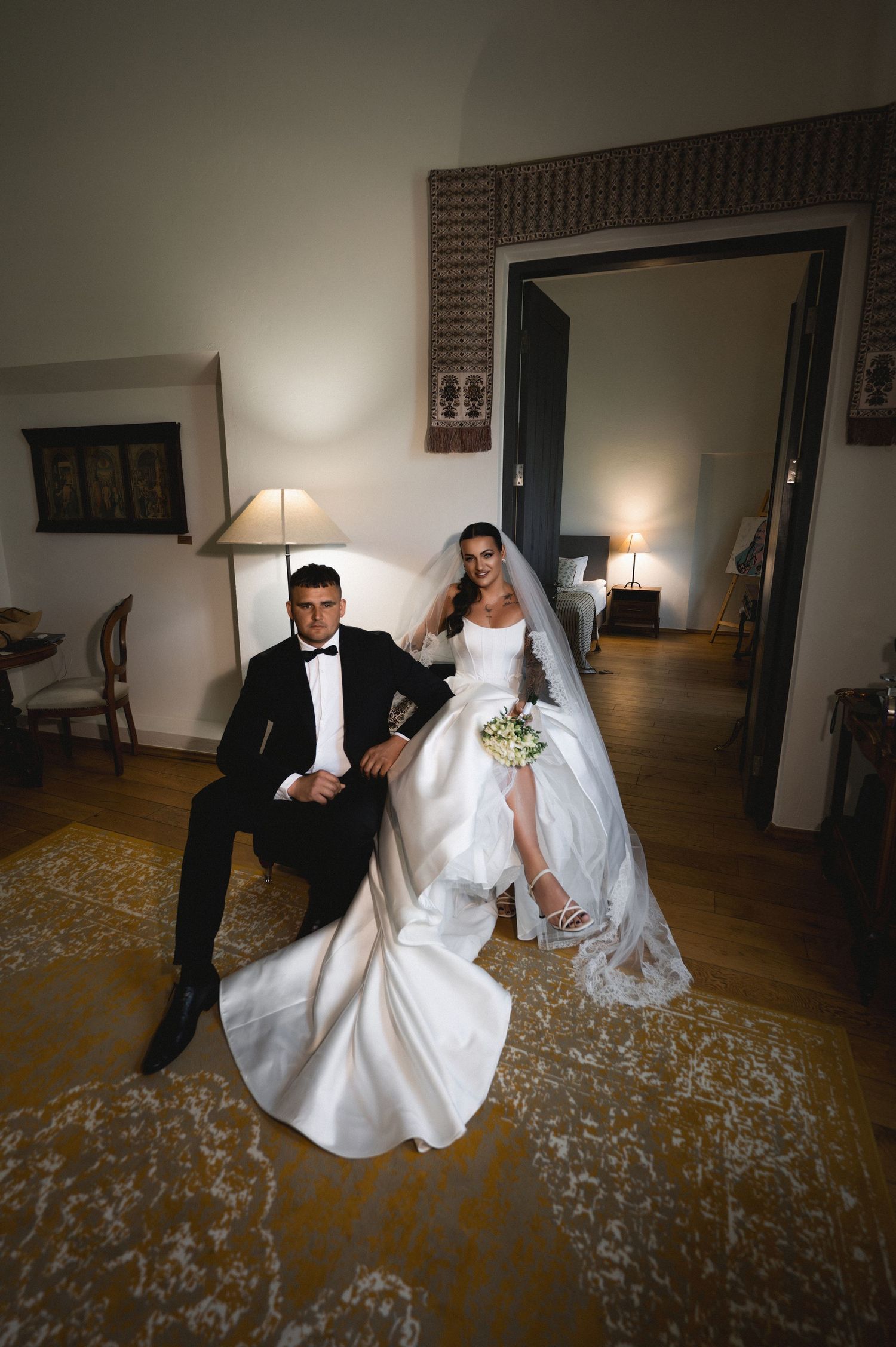 Bride in white gown with veil and groom in black tuxedo pose elegantly in a warmly lit room on their wedding day.