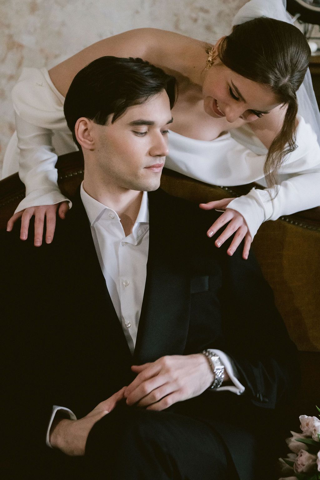 Elegant bride in white dress leans over groom in black suit seated on vintage chair in romantic wedding portrait.