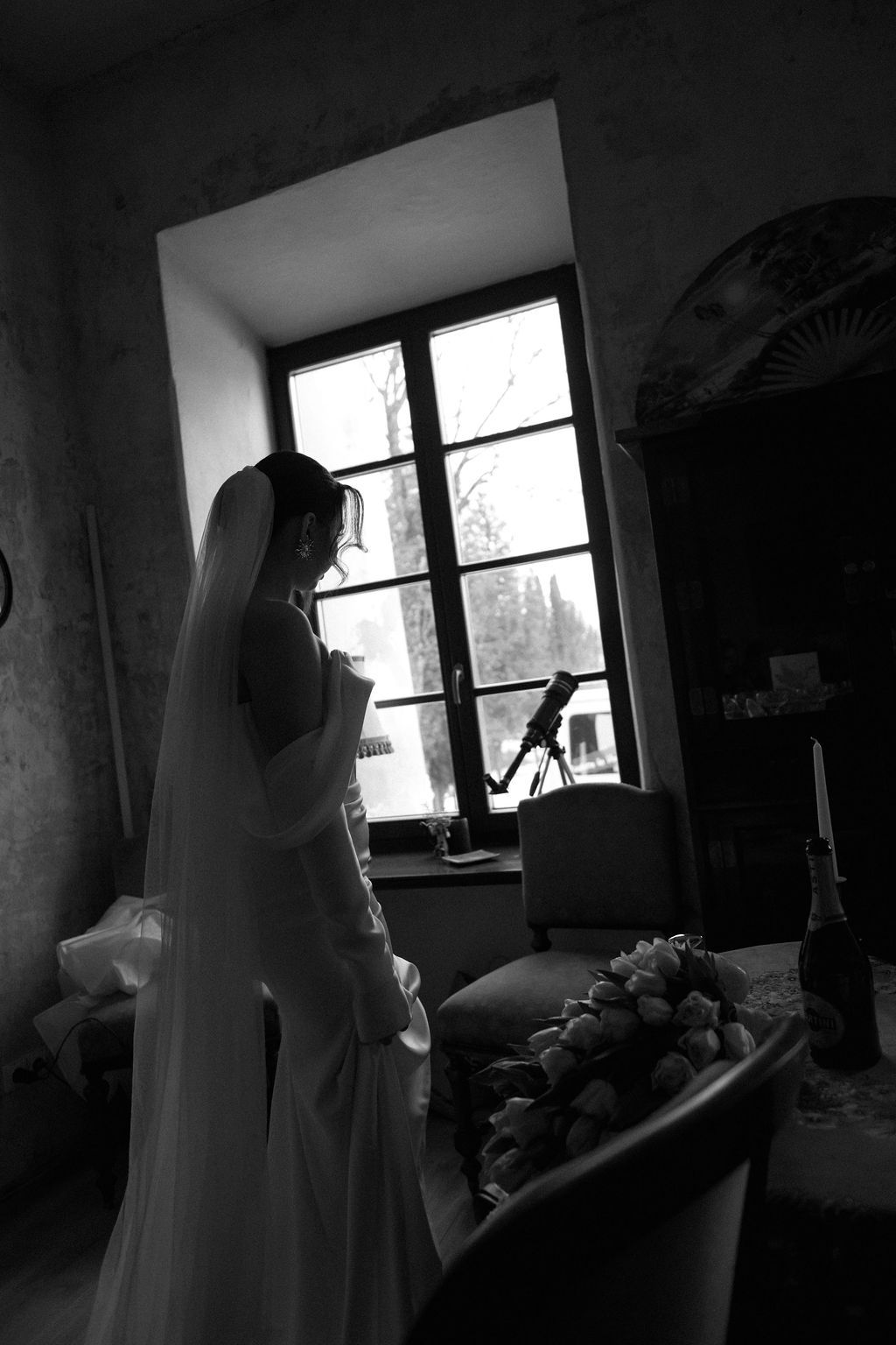 Black and white photo of a bride in wedding dress and veil standing by a window with bouquet nearby.
