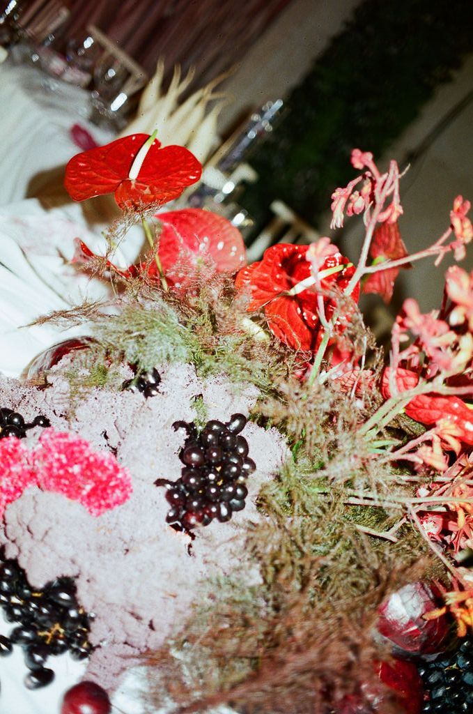 Red anthurium flowers and dark berries arranged in a lush floral centerpiece with green ferns and pink blooms.