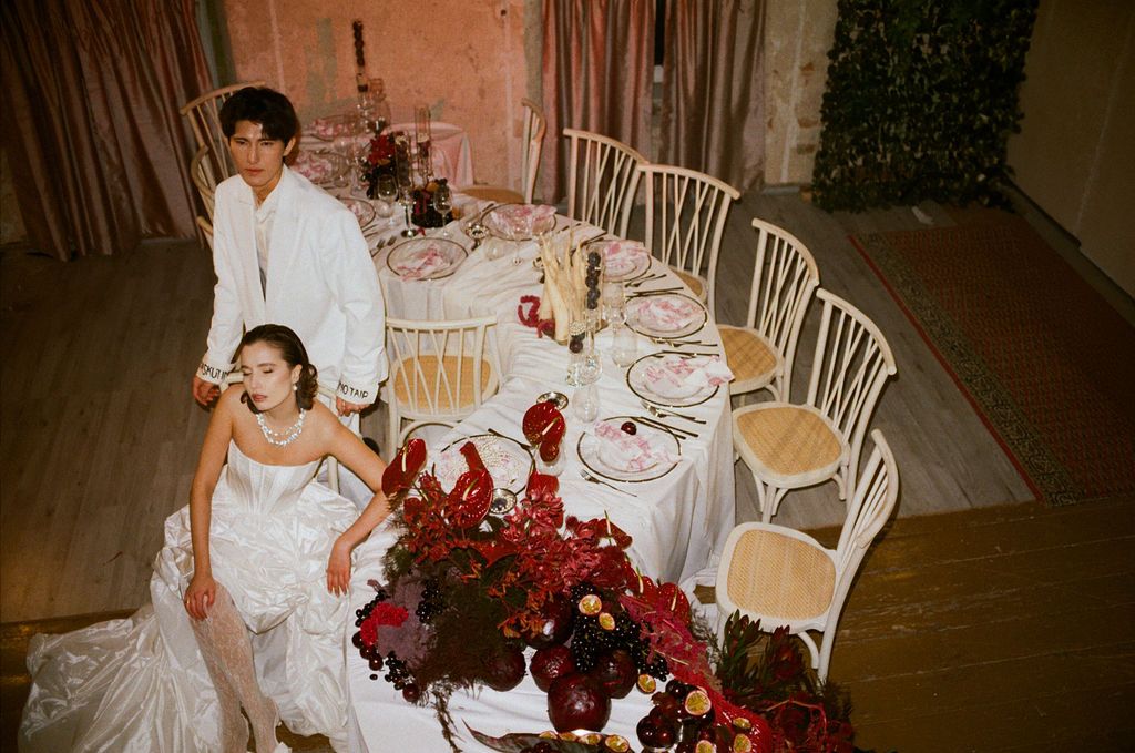 Bride in white gown and groom in white suit pose at elegant wedding reception table with red floral arrangements.