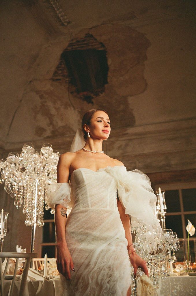 Bride in strapless white feather wedding gown with tulle sleeve poses near crystal candelabras in rustic venue.