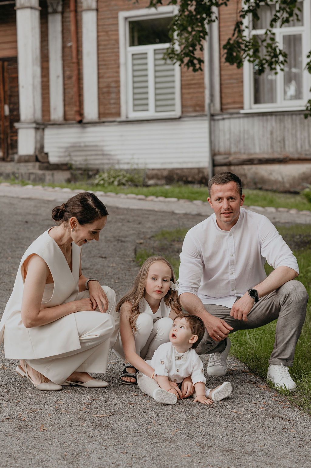 Family of four in white outfits posing outdoors near a brick building, parents with two young children on a sunny day.