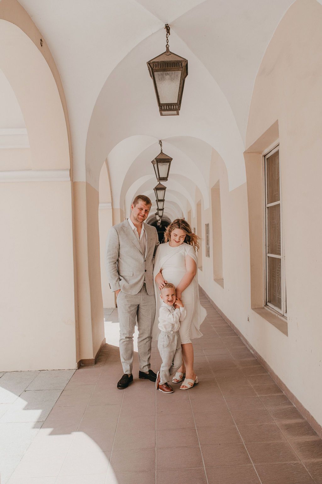 A family of three poses in an elegant arched corridor with hanging lanterns in a European-style building.