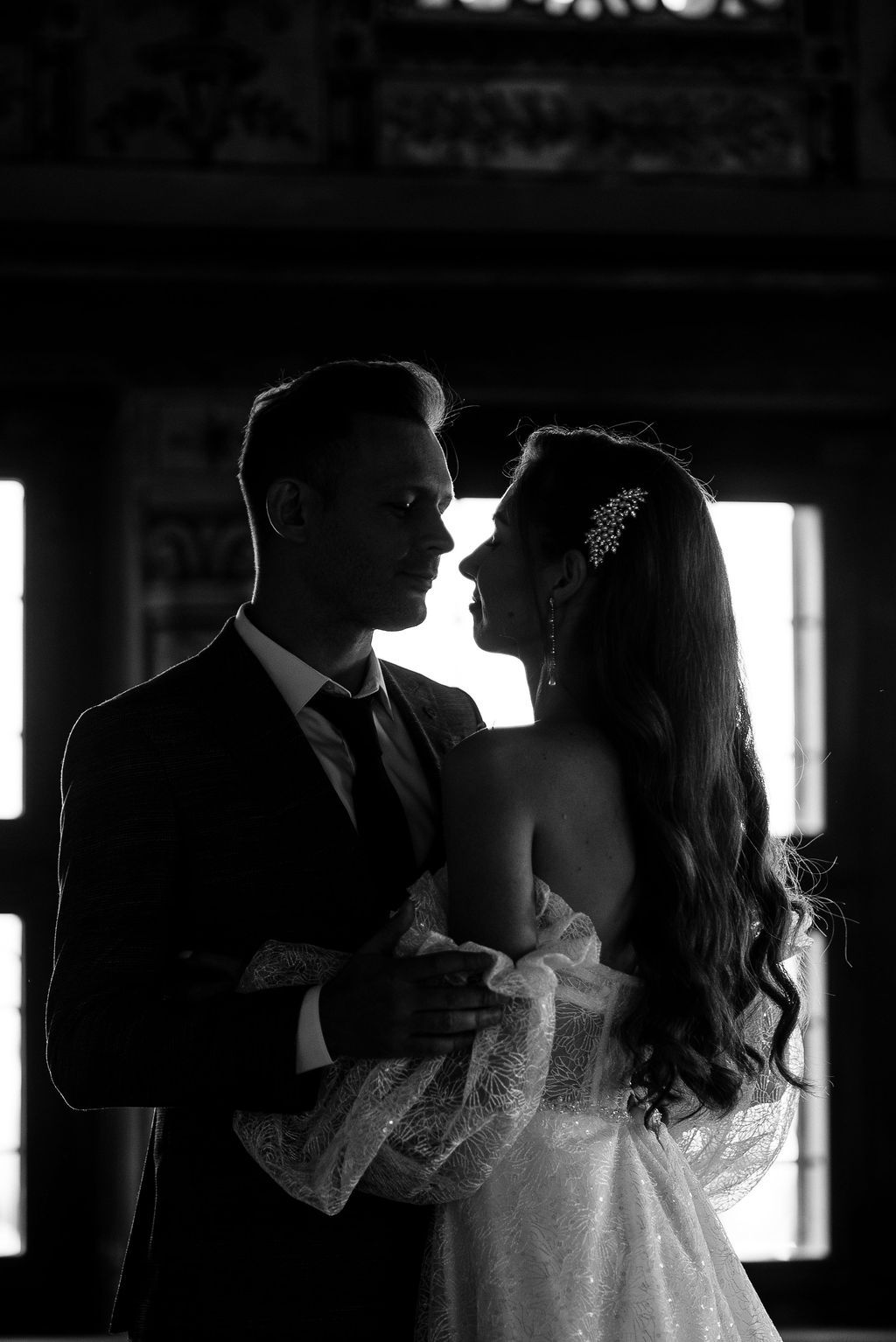 Black and white wedding photo of bride and groom sharing an intimate moment, backlit by windows.