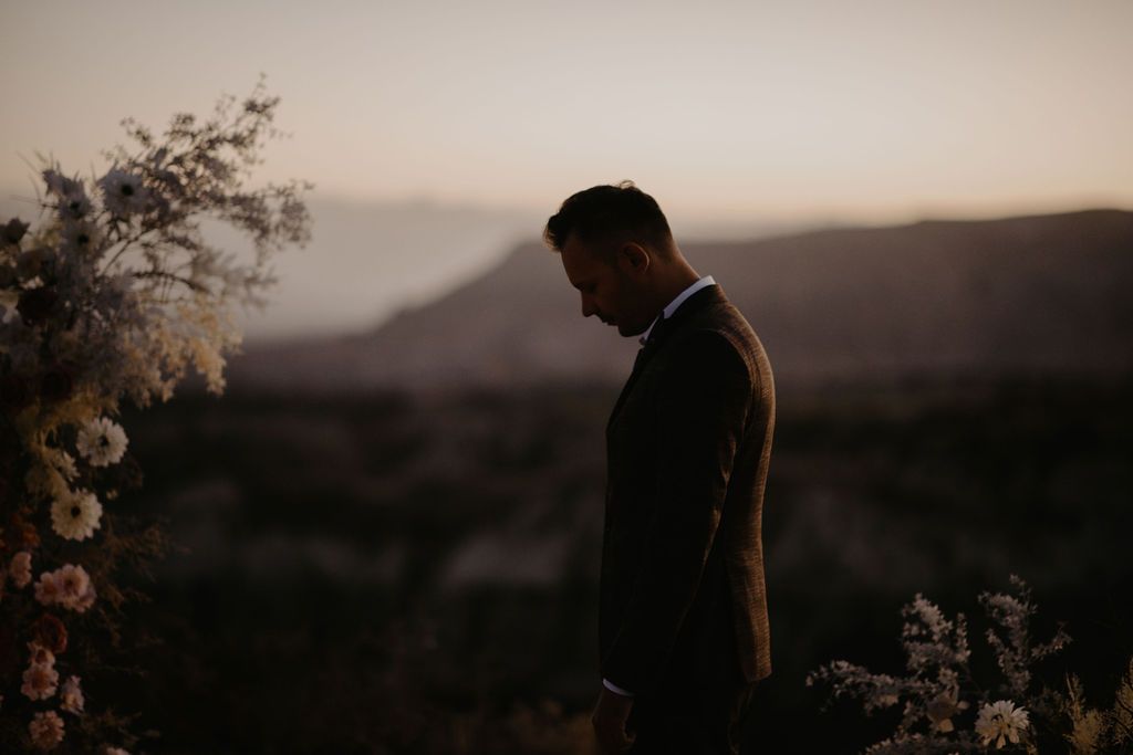 Groom in dark suit standing at outdoor wedding altar with floral arrangement at sunset with mountain backdrop.