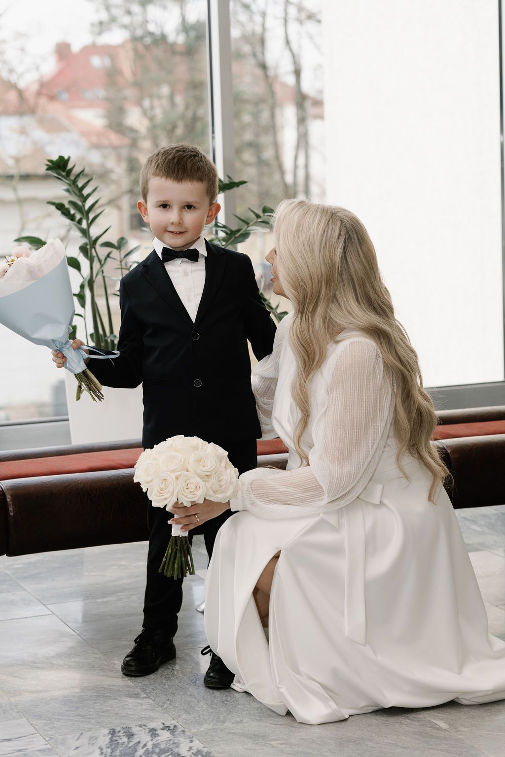 A young boy in a black tuxedo presents white flowers to a blonde bride in a white dress at a wedding.