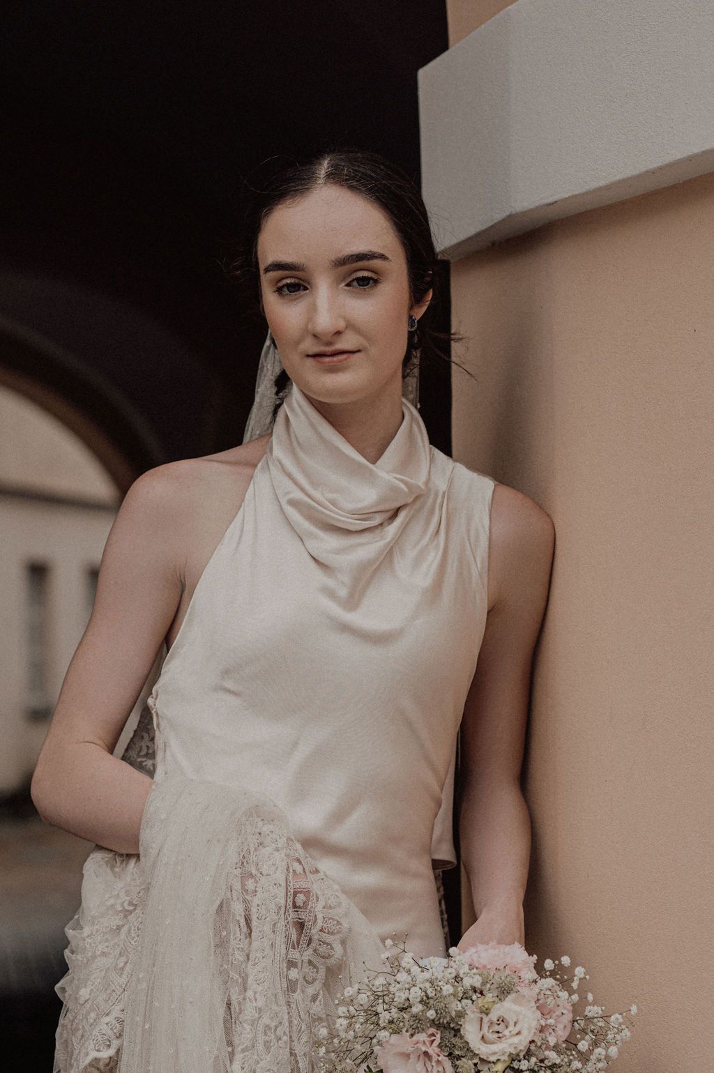 Elegant bride in ivory satin halter gown holding a delicate bouquet of white and pink flowers outdoors.