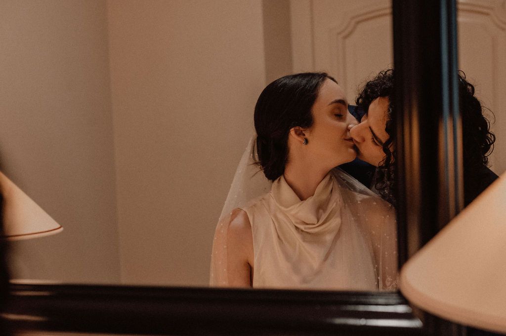 Two brides share a tender kiss in a softly lit elegant room on their wedding day.
