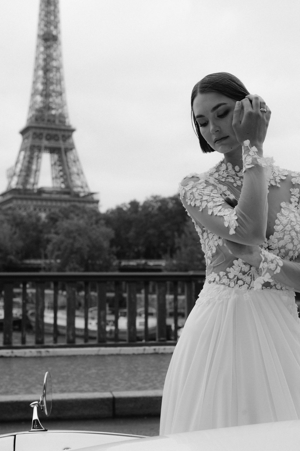 Bride in floral lace wedding gown poses near Eiffel Tower in elegant black and white Paris photo.