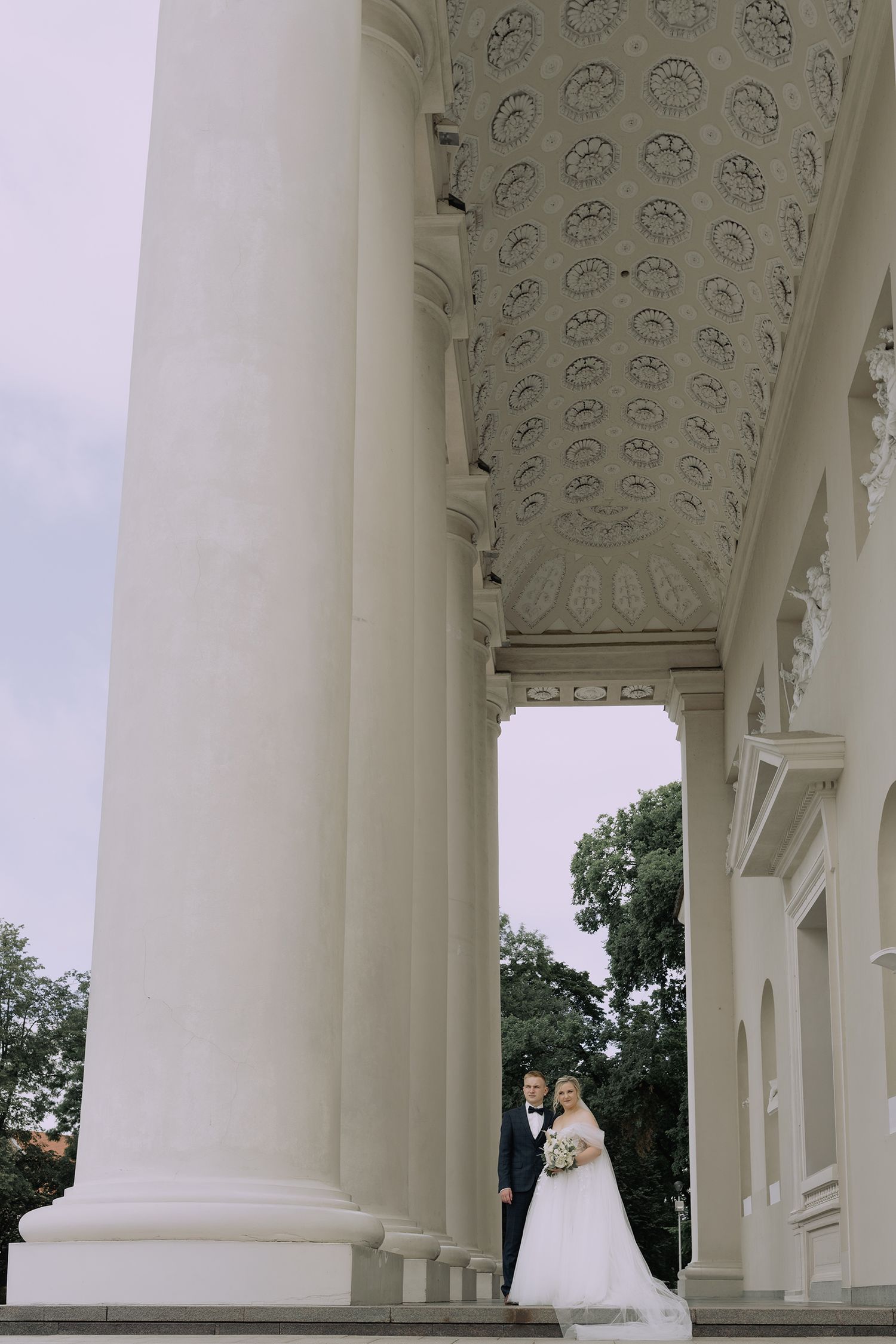 Elegant couple stands between white classical columns with ornate ceiling details at a grand historic venue.