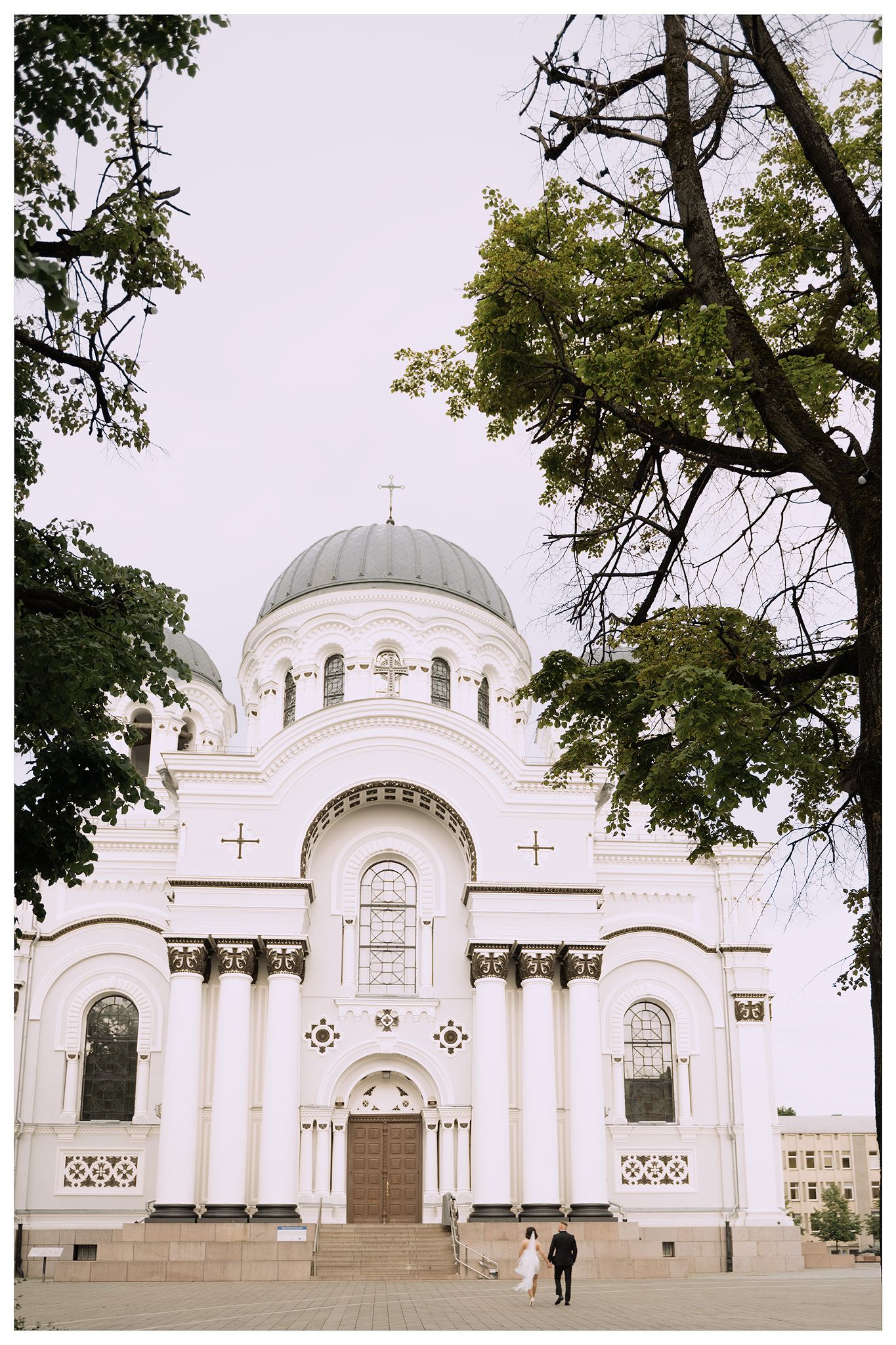 A panoramic view of a white Orthodox church with domed roofs and ornate architectural details framed by trees.