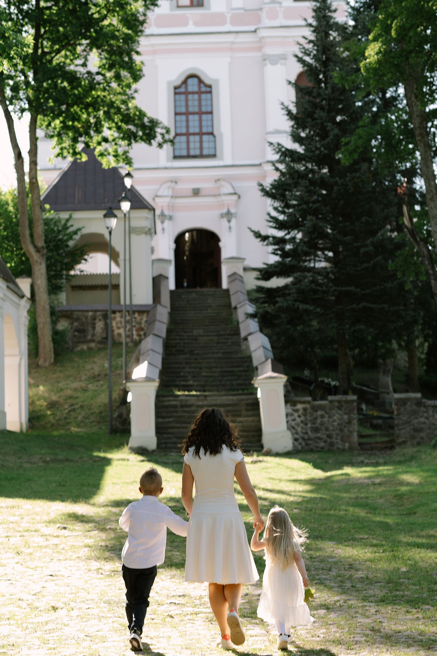 Three people walk up stone steps leading to a historic building surrounded by trees and greenery in a park setting.