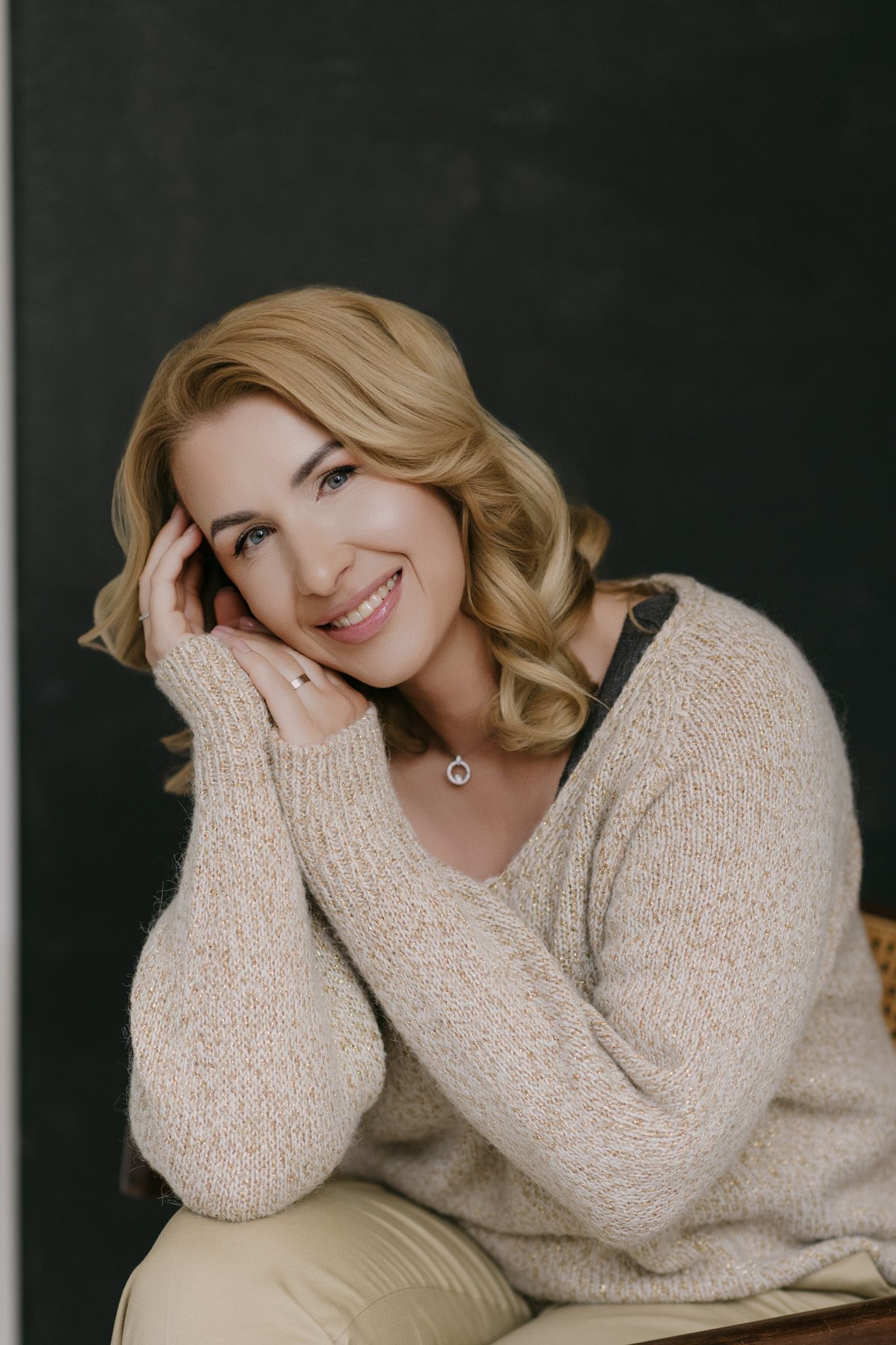 Smiling blonde woman in beige knit sweater posing with hands near face against dark background.