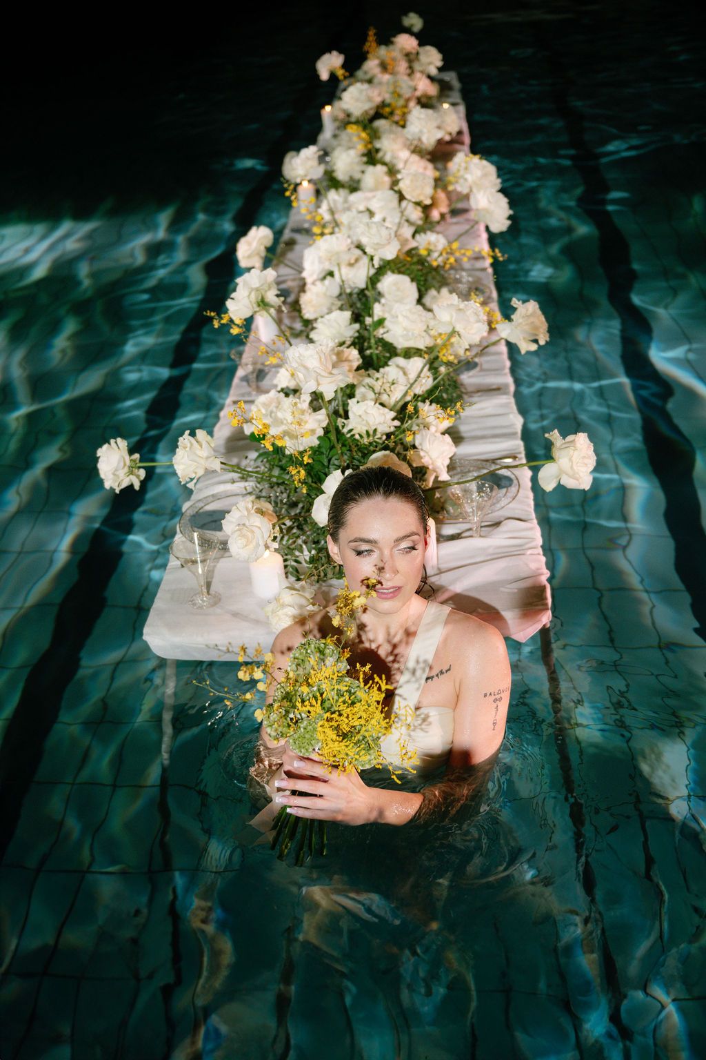 Woman standing in a pool holding a yellow flower bouquet beside a floating table covered in white roses and blooms.
