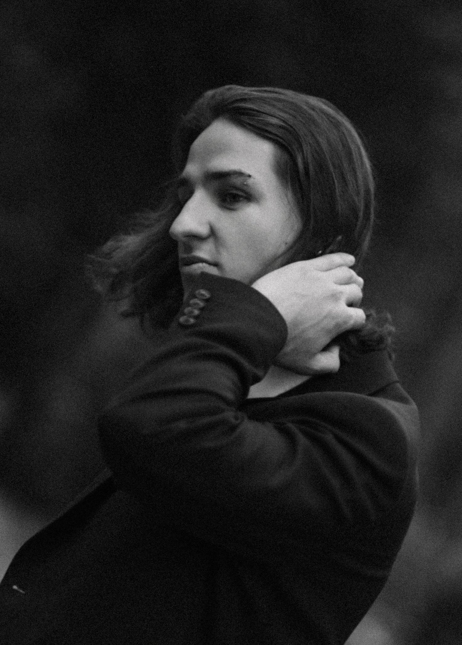Black and white portrait of a person with shoulder-length dark hair, wearing a dark blazer, hand raised to the neck, gazing sideways.
