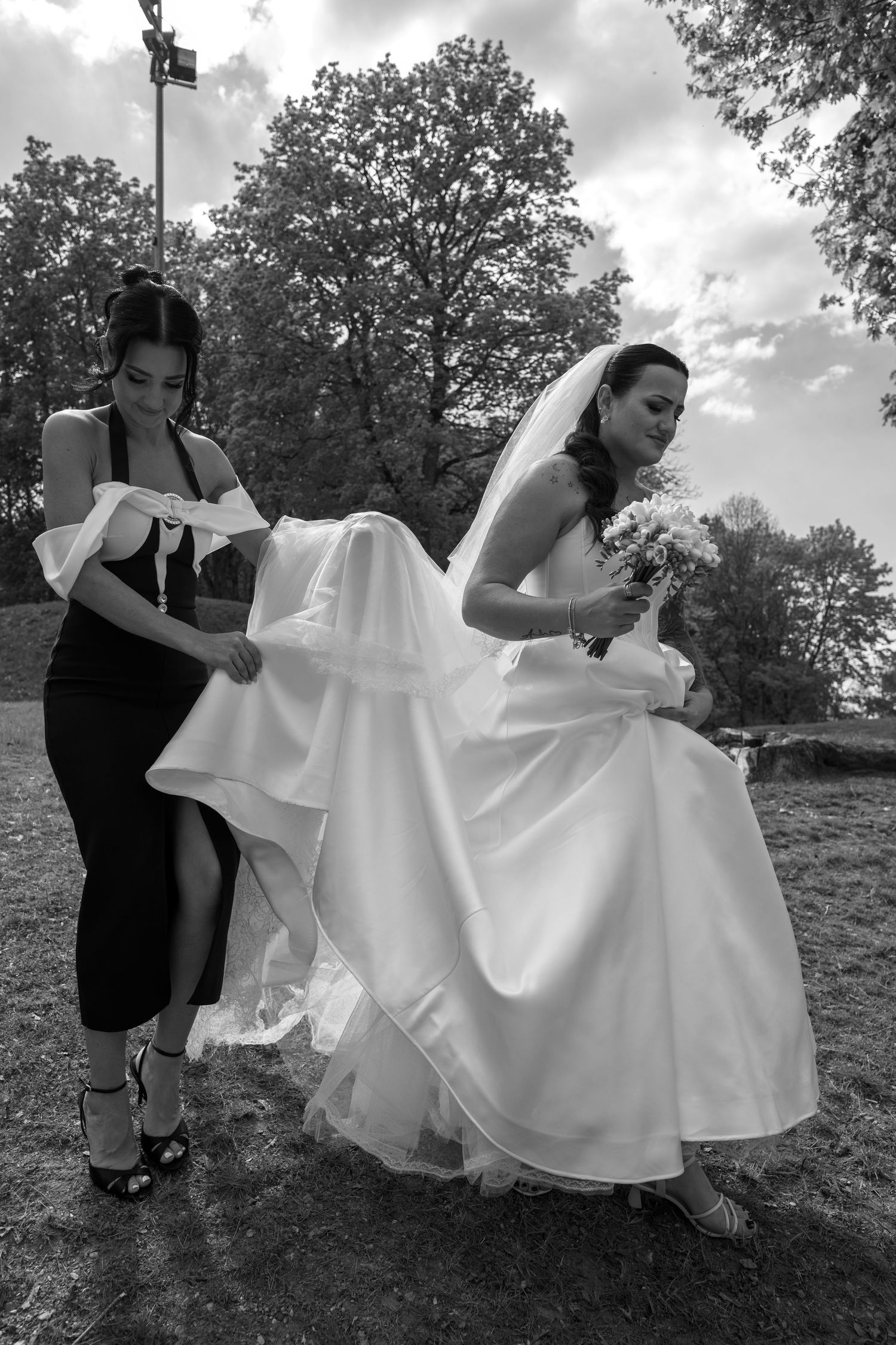 A bridesmaid in a black dress lifts the train of a bride's white wedding gown outdoors beneath tall trees in black and white.