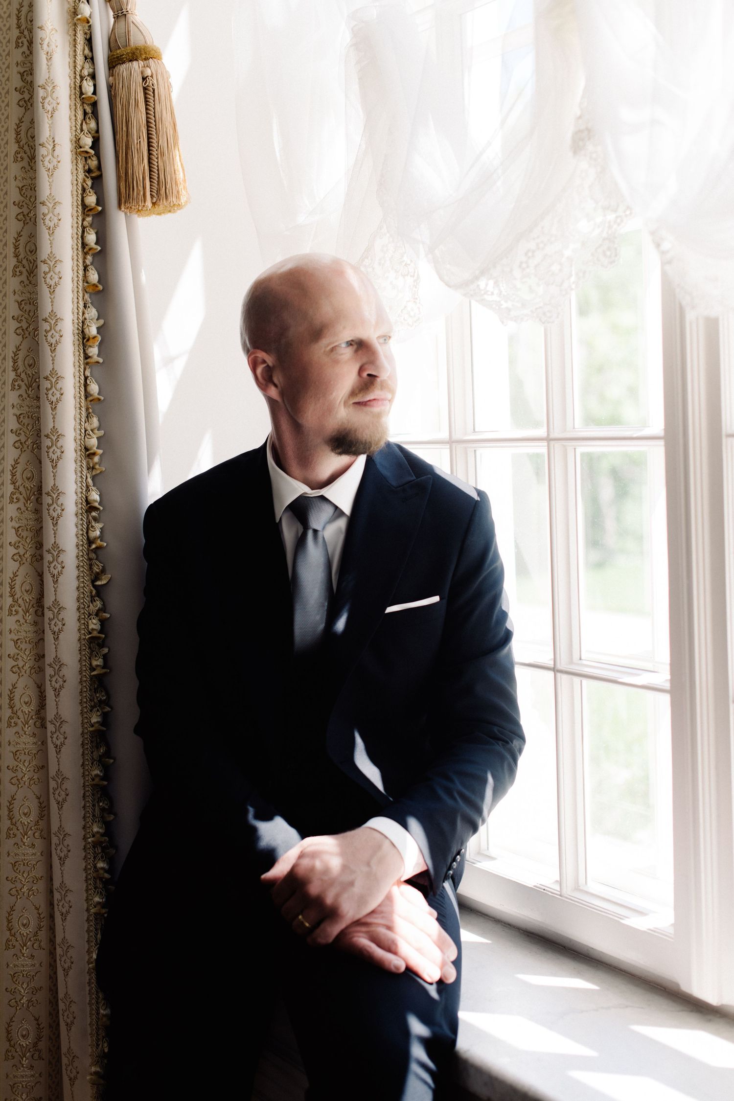 Smiling groom in a navy suit and gray tie sits by a bright window with sheer curtains, gazing into the distance.