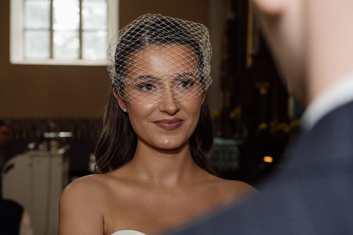 Smiling bride in a strapless white dress and birdcage veil stands during a wedding ceremony, with a groom blurred in the foreground.