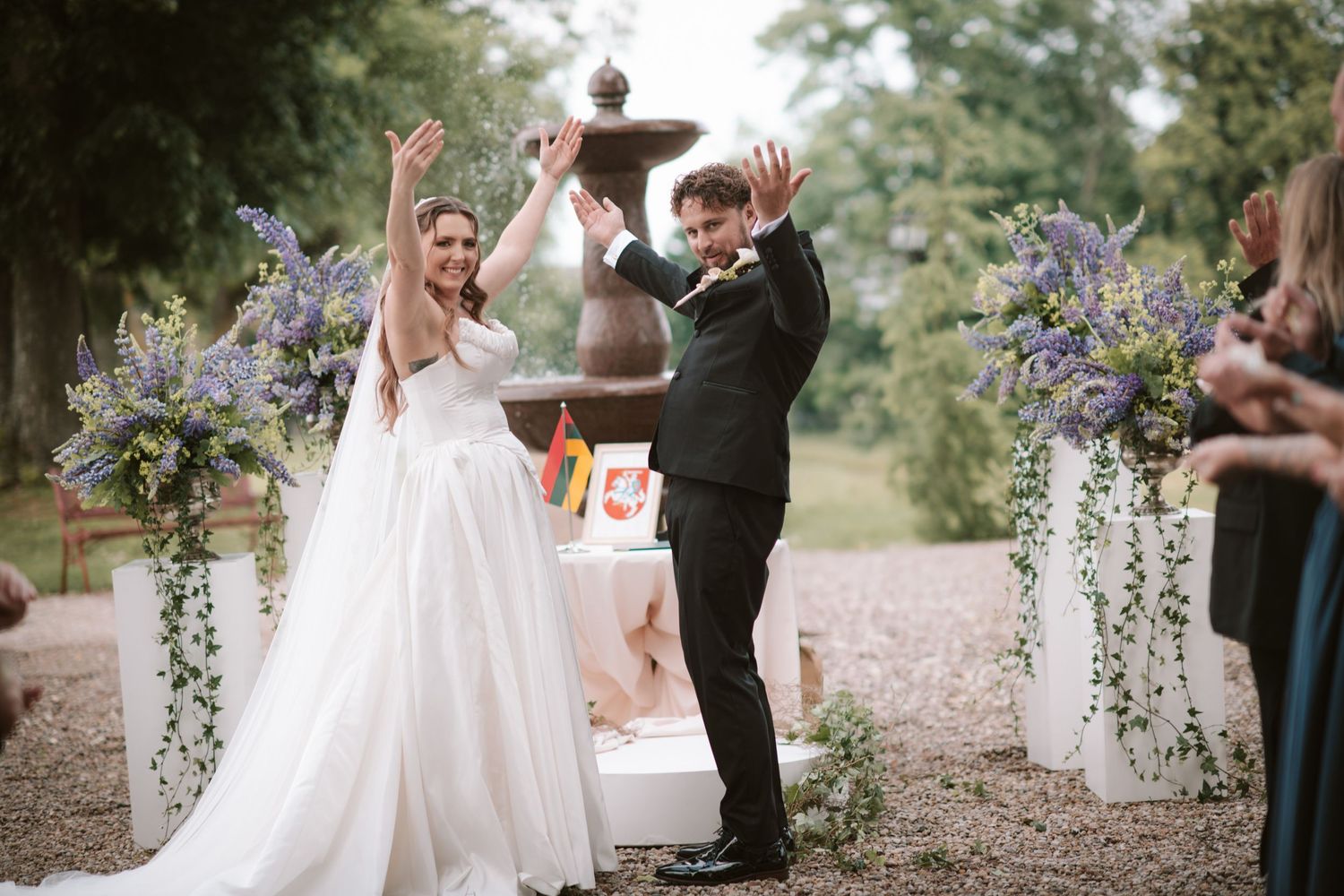 Joyful bride and groom raising arms in celebration at outdoor wedding ceremony with purple floral arrangements.