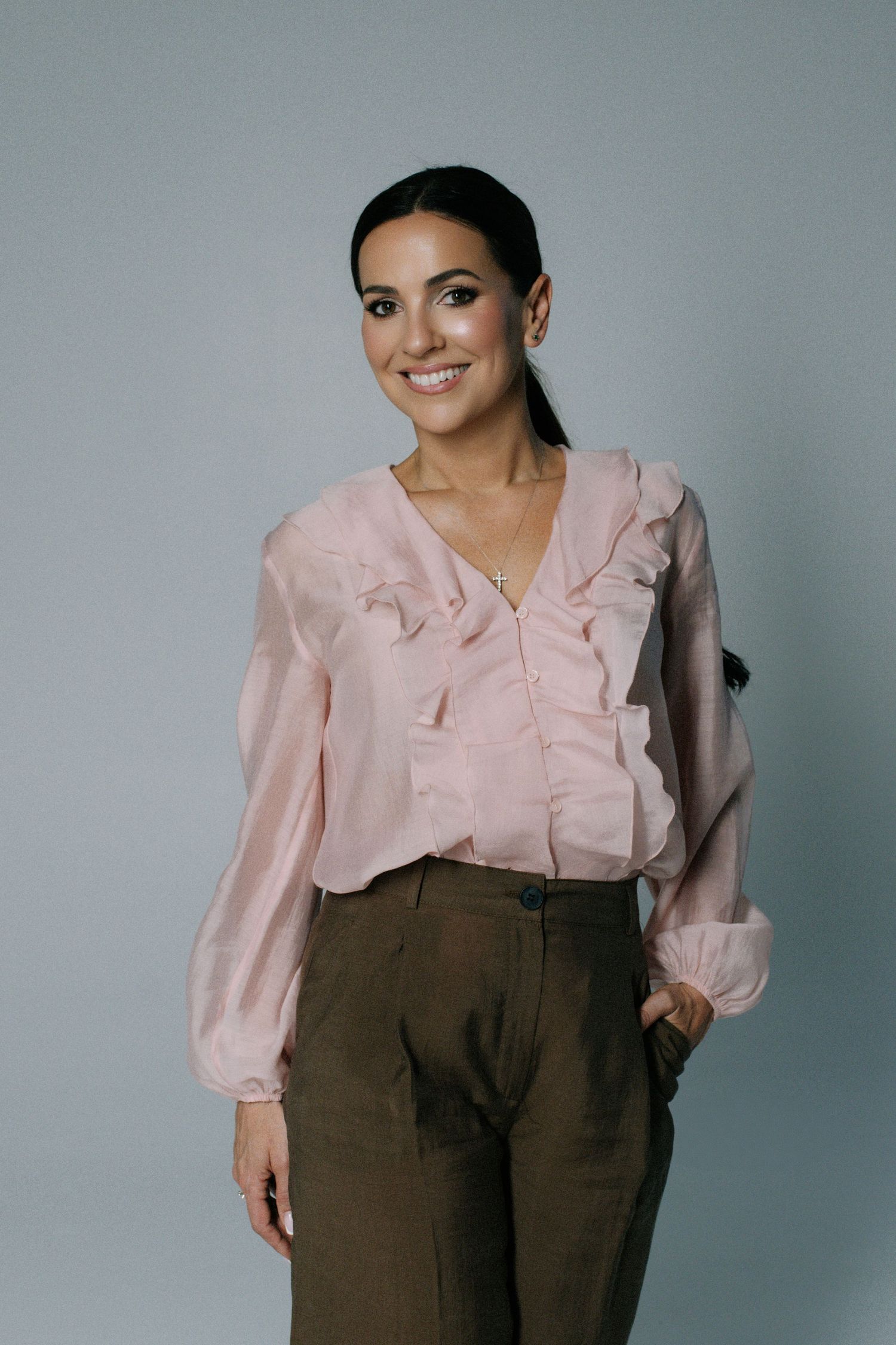 Woman in pink ruffled blouse and olive trousers smiling against gray background.