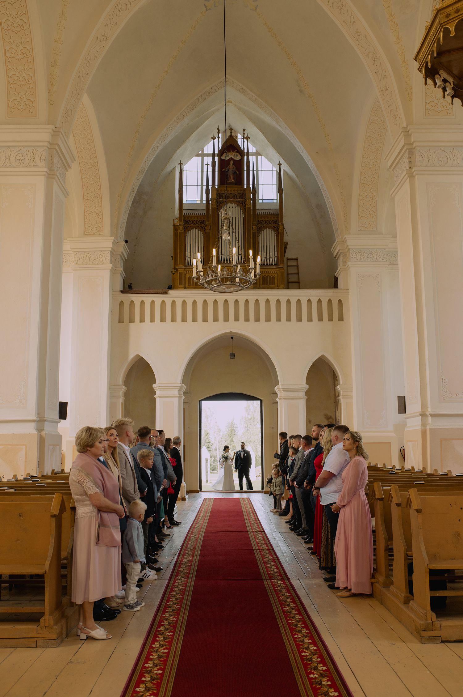 A church interior with a red carpet aisle and white arched walls leading to an altar with guests lining the sides.