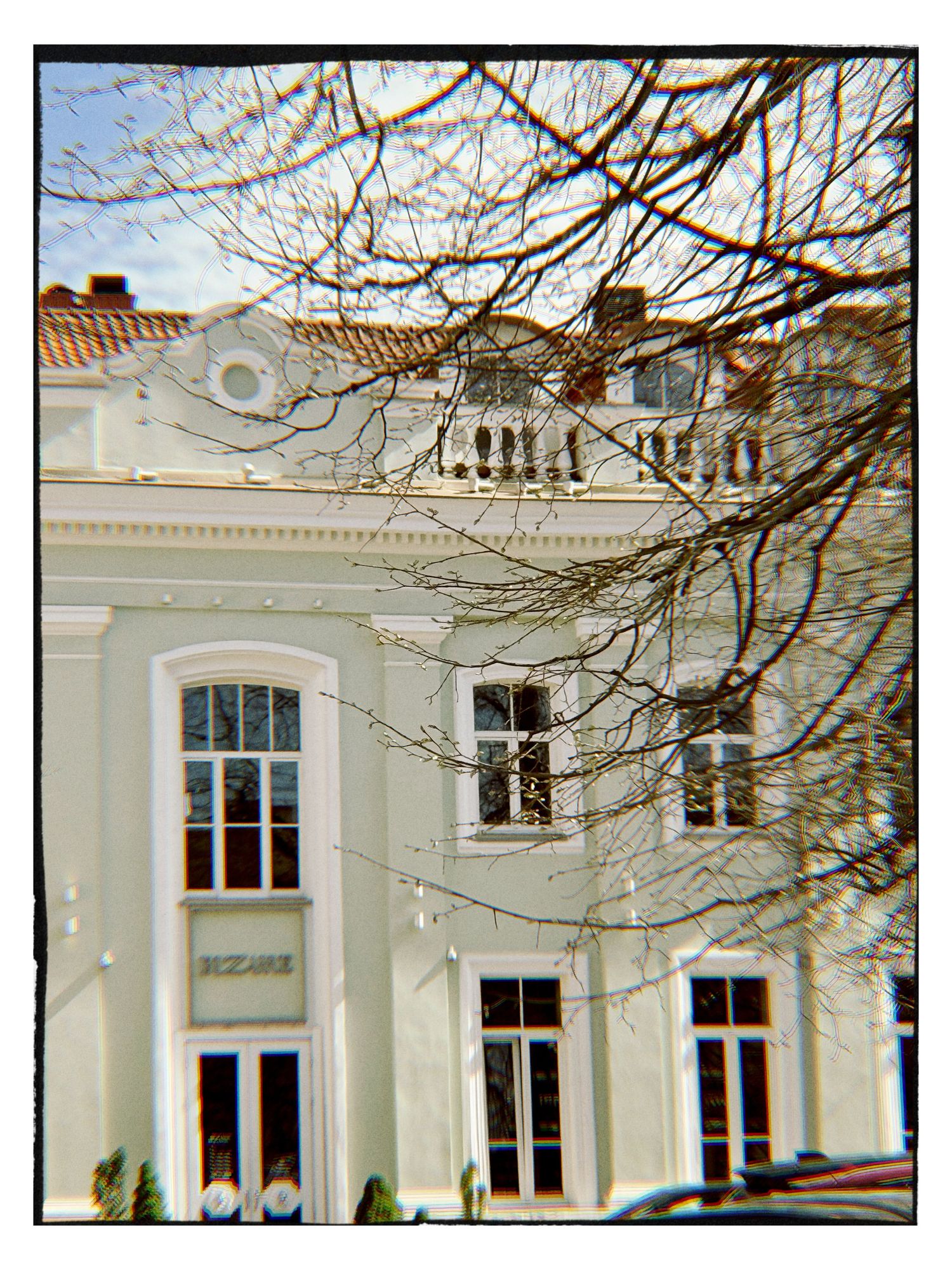 Historic white neoclassical building with arched windows and ornate balcony, framed by bare winter trees against a bright sky.